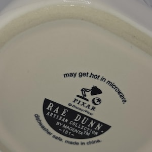 May include: White ceramic mug with the text "LAN" and the words "may get hot in microwave" printed on the bottom. The mug features the Pixar logo and the Rae Dunn Artisan Collection logo.
