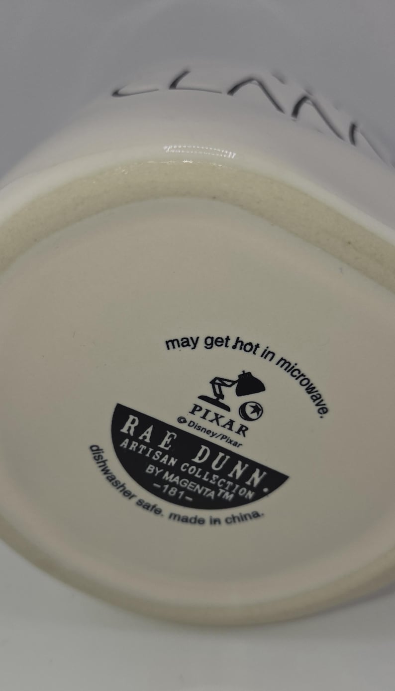 May include: White ceramic mug with the text "LAN" and the words "may get hot in microwave" printed on the bottom. The mug features the Pixar logo and the Rae Dunn Artisan Collection logo.