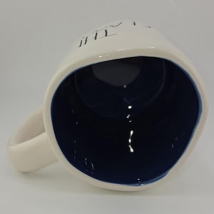 May include: White ceramic mug with a navy blue interior and a curved handle. The mug has the word "THE" printed on the side. The mug is angled, showing the inside and handle.