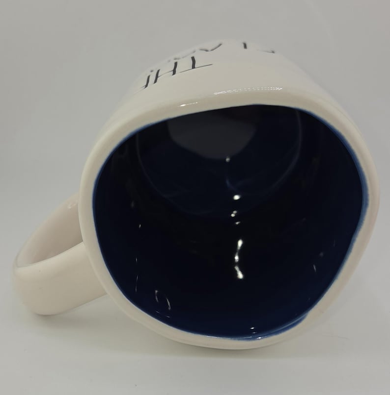 May include: White ceramic mug with a navy blue interior and a curved handle. The mug has the word "THE" printed on the side. The mug is angled, showing the inside and handle.