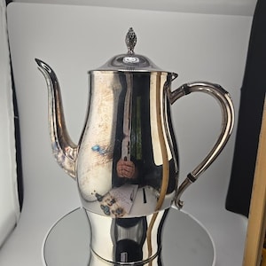 May include: A silver-colored teapot with a curved spout and handle. The teapot has a lid with a decorative finial. The teapot is sitting on a mirrored surface. The teapot is likely made of metal.