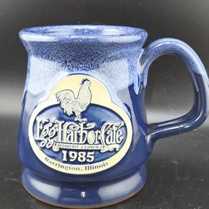 May include: A deep blue ceramic mug with a glossy finish and a lighter blue drip glaze at the rim. The front features an embossed oval emblem with a rooster illustration, the text Egg Harbor Cafe, Breakfast & Lunch, 1985, and Barrington, Illinois.