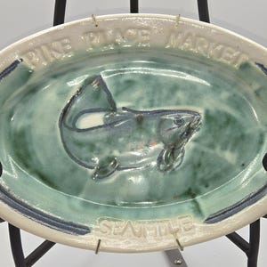 May include: Oval ceramic dish with a raised fish design in the center. The dish has a light green interior and a cream-colored rim with the words "Pike Place Market" and "Seattle" embossed around the edge. A dark blue border accents the rim.