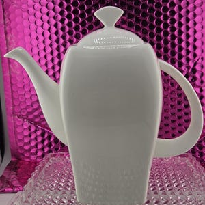 May include: A white ceramic teapot with a curved spout and handle. The teapot has a lid with a small finial. The teapot is sitting on a clear, textured glass surface. The background is a shiny, pink surface.