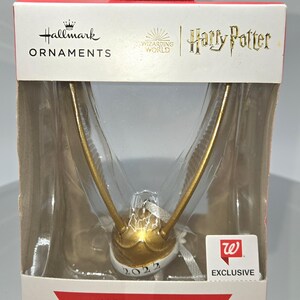 May include: A Harry Potter Christmas tree ornament featuring a golden Snitch with outstretched wings. The ornament is in a clear plastic case with the Hallmark and Wizarding World logos. The year "2022" is visible on the Snitch.