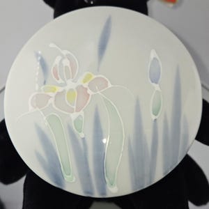 May include: A round, white ceramic plate with a floral design. The design features a stylized iris with pink, yellow, and green accents, surrounded by blue and green leaf-like shapes. The plate has a glossy finish.