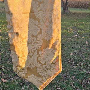 May include: Gold-colored damask table runner with a floral pattern. The runner has a decorative tassel at the end. The fabric is illuminated by sunlight, creating a warm, inviting aesthetic. The runner is displayed outdoors.