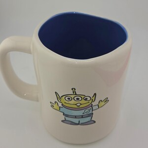 May include: Cream-colored ceramic mug with a blue interior and a curved handle. The mug features a cartoon illustration of a green alien with three eyes, wearing a light blue uniform. The mug is designed for beverages.