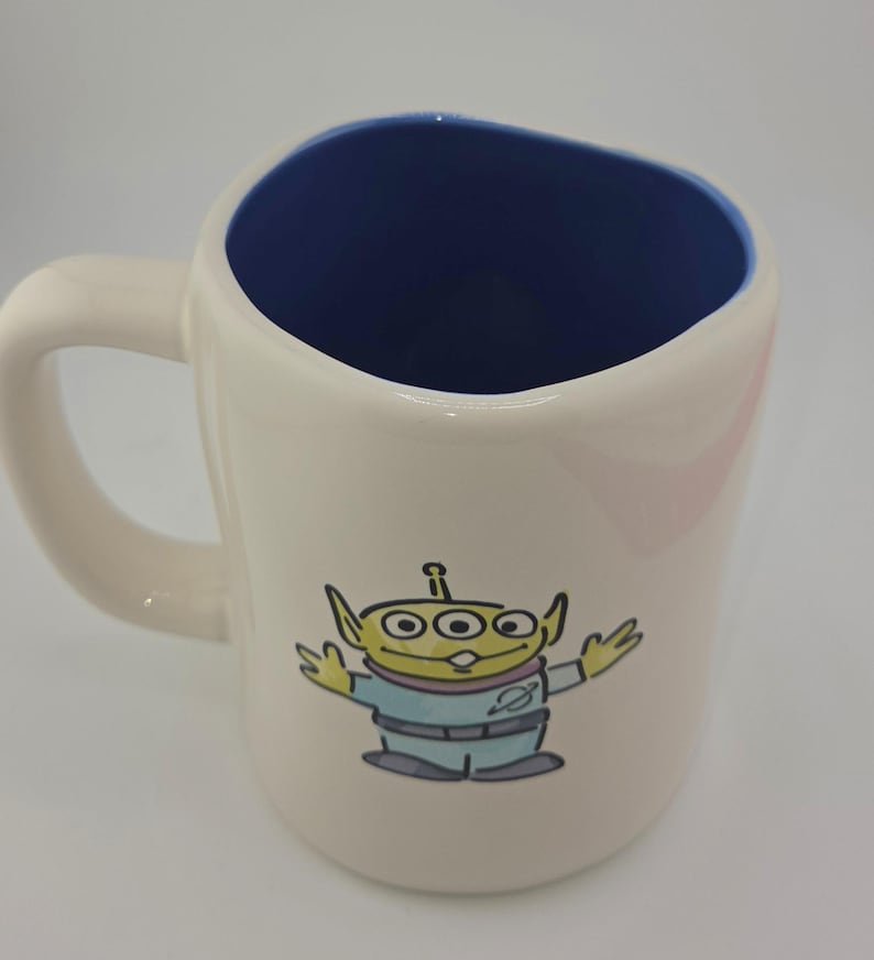 May include: Cream-colored ceramic mug with a blue interior and a curved handle. The mug features a cartoon illustration of a green alien with three eyes, wearing a light blue uniform. The mug is designed for beverages.
