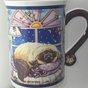May include: A ceramic mug featuring a sleeping cat illustration. The mug has a dark brown handle with a mouse design. The artwork includes a window scene with stars, a lamp, and the text "GARY PATTERSON".