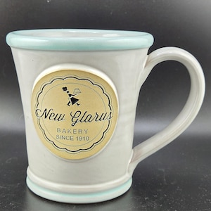 May include: A light gray ceramic mug with a light blue rim and base. A circular tan emblem on the front features a black silhouette of a baker holding a cake, the text New Glarus, and the words BAKERY SINCE 1910 in a decorative black border.