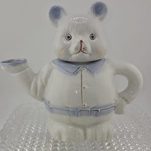 May include: A white ceramic teapot in the shape of a bear with blue accents on the ears, collar, and belt. The teapot has a spout, handle, and a lid that forms the bear's head. The bear's face has painted details.