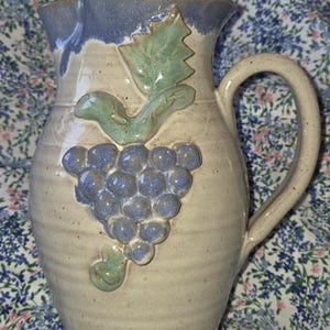 May include: A handmade ceramic pitcher with a light beige body, a blue rim, and a large handle. The pitcher is decorated with a cluster of blue grapes and green leaves.