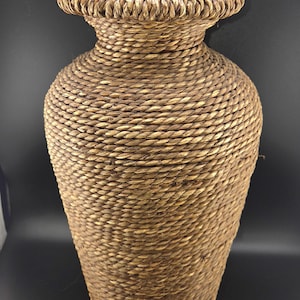 May include: A tall, decorative vase woven from natural brown fibers. The vase has a wide, flared rim and a rounded body that tapers towards the base. The texture is rough and the color is a warm, earthy brown.