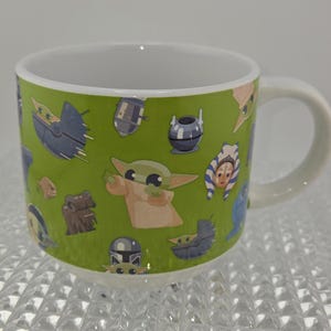 Może przedstawiać: Biały ceramiczny kubek z zielonym tłem z postaciami z kresk&oacute;wek z popularnej serii science fiction. Kubek ma biały uchwyt i białe wnętrze. Postacie obejmują małego zielonego kosmitę, statki kosmiczne i inne postacie.