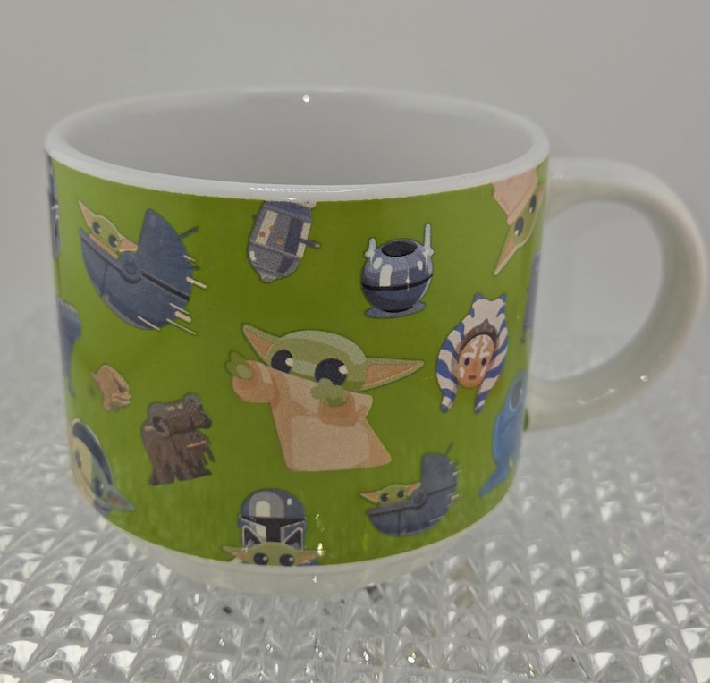 Może przedstawiać: Biały ceramiczny kubek z zielonym tłem z postaciami z kresk&oacute;wek z popularnej serii science fiction. Kubek ma biały uchwyt i białe wnętrze. Postacie obejmują małego zielonego kosmitę, statki kosmiczne i inne postacie.