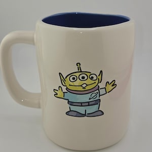 May include: Cream-colored ceramic mug with a blue interior. The mug features a cartoon illustration of a green alien with three eyes, wearing a blue uniform. The mug has a curved handle.