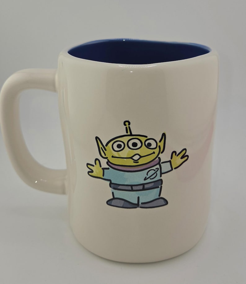 May include: Cream-colored ceramic mug with a blue interior. The mug features a cartoon illustration of a green alien with three eyes, wearing a blue uniform. The mug has a curved handle.