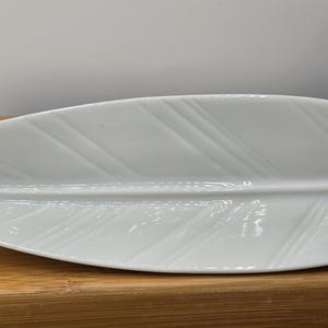 May include: A white, leaf-shaped ceramic serving dish. The dish has a long, slender shape with a textured surface, mimicking the veins of a leaf. It rests on a wooden tray, suggesting a decorative or serving purpose.