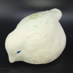 May include: A ceramic bird figurine in a soft, off-white color. The bird has a rounded body and a small, pointed beak. A small blue accent marks the eye. The surface has a textured, slightly rough appearance.