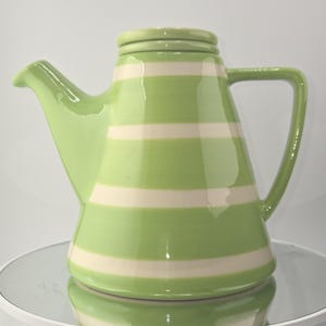 May include: A ceramic teapot with a light green and cream horizontal striped pattern. The teapot has a spout, a handle, and a lid. The teapot is sitting on a white surface.