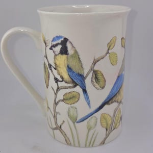 May include: White ceramic mug featuring a detailed illustration of two blue and yellow birds perched on branches with green leaves. The mug has a curved handle and a smooth, glossy finish, perfect for enjoying hot beverages.