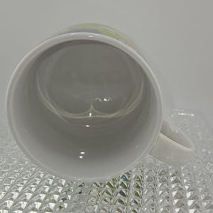 Może przedstawiać: Biały ceramiczny kubek z uchwytem, ​​widziany z g&oacute;ry. Wnętrze kubka jest jasno szare. Kubek stoi na przezroczystej, teksturowanej powierzchni.