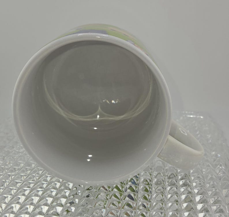 Może przedstawiać: Biały ceramiczny kubek z uchwytem, ​​widziany z g&oacute;ry. Wnętrze kubka jest jasno szare. Kubek stoi na przezroczystej, teksturowanej powierzchni.