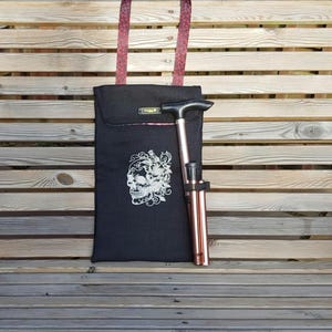 May include: A black tote bag featuring a white skull design and a copper-coloured folding cane. The bag has burgundy straps and a small decorative label. The cane is folded and rests against the bag, placed on a wooden bench.