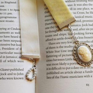 May include: Two white satin ribbon bookmarks with silver metal accents. One bookmark has a pearl charm and the other has a silver cameo charm with a white cameo portrait. The bookmarks are on a book with text about the Titanic musicians.