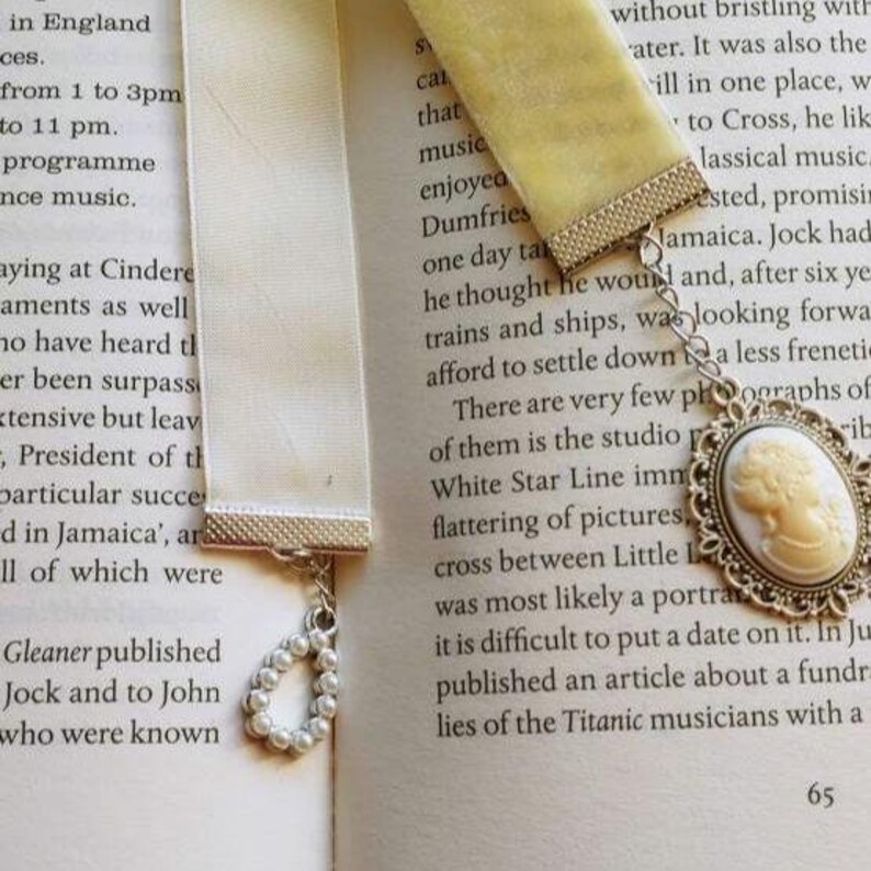 May include: Two white satin ribbon bookmarks with silver metal accents. One bookmark has a pearl charm and the other has a silver cameo charm with a white cameo portrait. The bookmarks are on a book with text about the Titanic musicians.