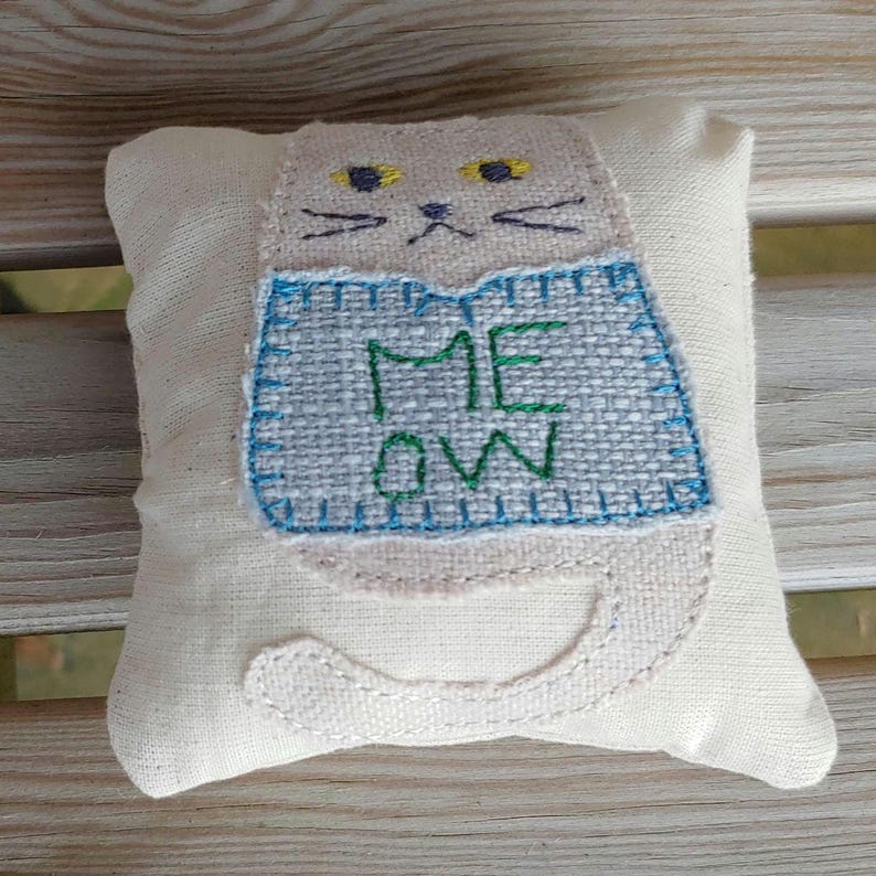 May include: A white fabric cat pillow with embroidered yellow eyes, a black nose, and whiskers. The cat is wearing a blue and gray vest with the word "MEOW" embroidered in green.