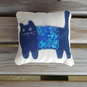 May include: A handmade blue cat pillow with a blue and green floral patterned fabric on the back. The pillow is made of a white fabric and has a simple design.