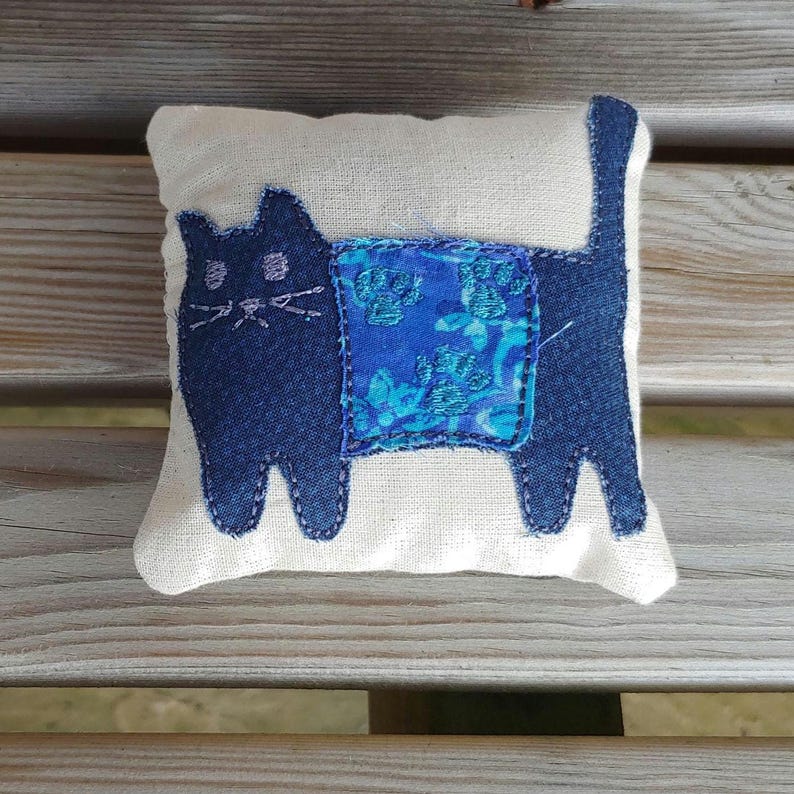 May include: A handmade blue cat pillow with a blue and green floral patterned fabric on the back. The pillow is made of a white fabric and has a simple design.