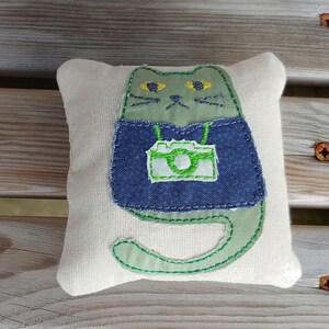 May include: A green cat with yellow eyes wearing a blue denim vest with a green camera embroidered on it. The cat is sitting on a white pillow.