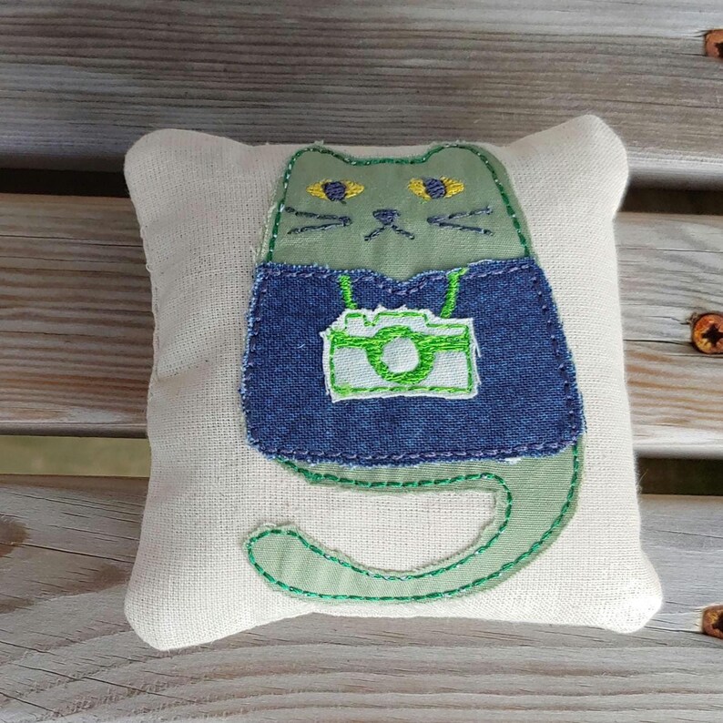 May include: A green cat with yellow eyes wearing a blue denim vest with a green camera embroidered on it. The cat is sitting on a white pillow.