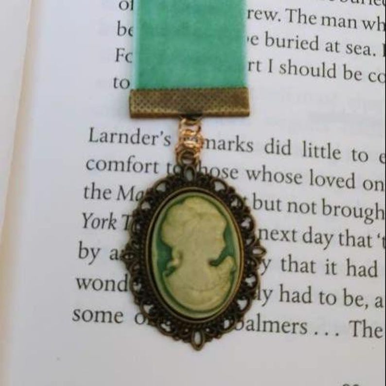 May include: A green velvet ribbon bookmark with a gold metal chain and a cameo pendant. The cameo is oval shaped and features a white silhouette of a woman's profile against a green background.