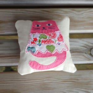 May include: A pink cat pillow with a floral print dress. The pillow is made of white fabric and has pink stitching.
