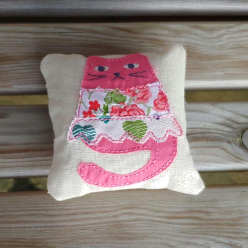 May include: A pink cat pillow with a floral print dress. The pillow is made of white fabric and has pink stitching.