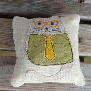 May include: A white fabric cat pillow with a green shirt, yellow tie, and glasses. The cat is embroidered with a simple face and whiskers.