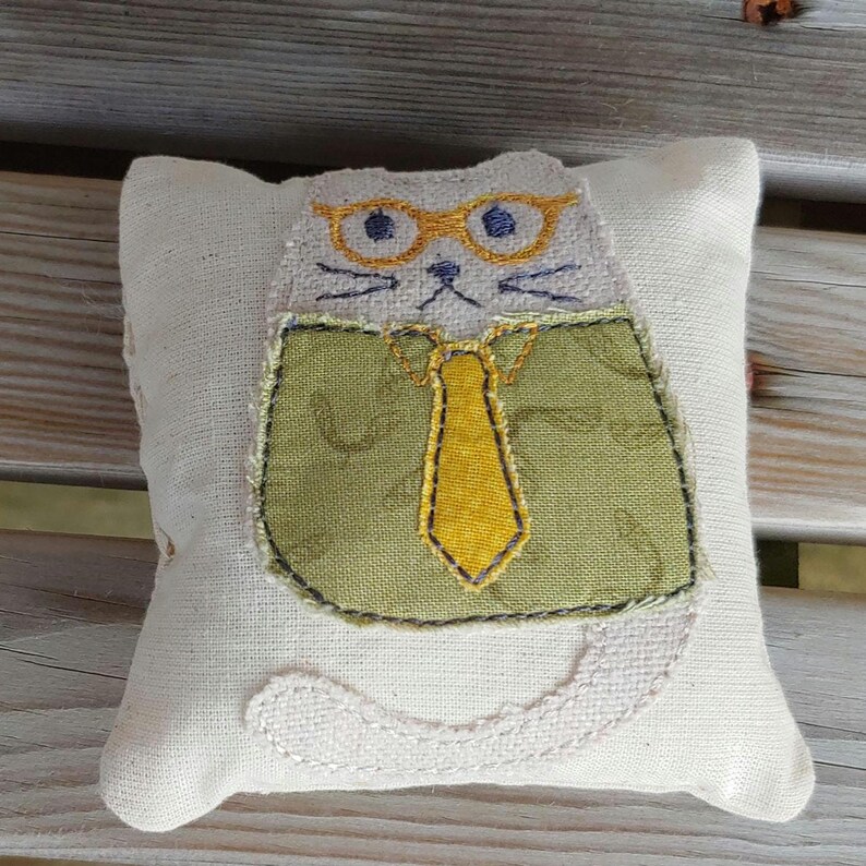 May include: A white fabric cat pillow with a green shirt, yellow tie, and glasses. The cat is embroidered with a simple face and whiskers.