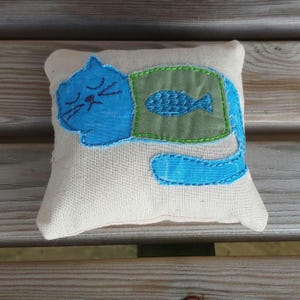 May include: A blue cat with a green fish on its chest, embroidered on a white pillow.