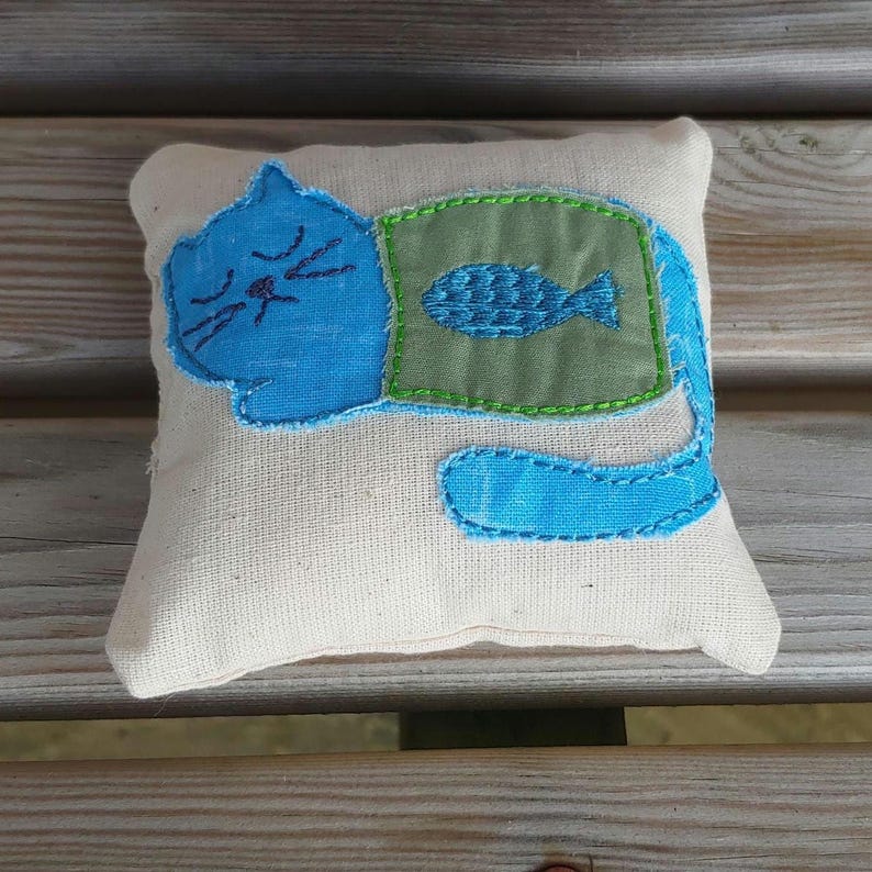 May include: A blue cat with a green fish on its chest, embroidered on a white pillow.