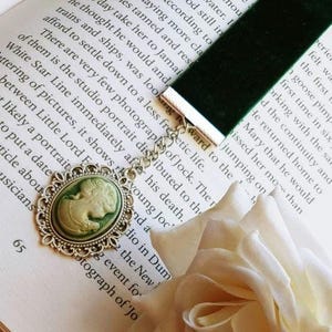 May include: A green velvet bookmark with a silver chain and a cameo pendant. The cameo is oval shaped and features a white porcelain image of a woman with a green background. The bookmark is lying on a book with a white rose.