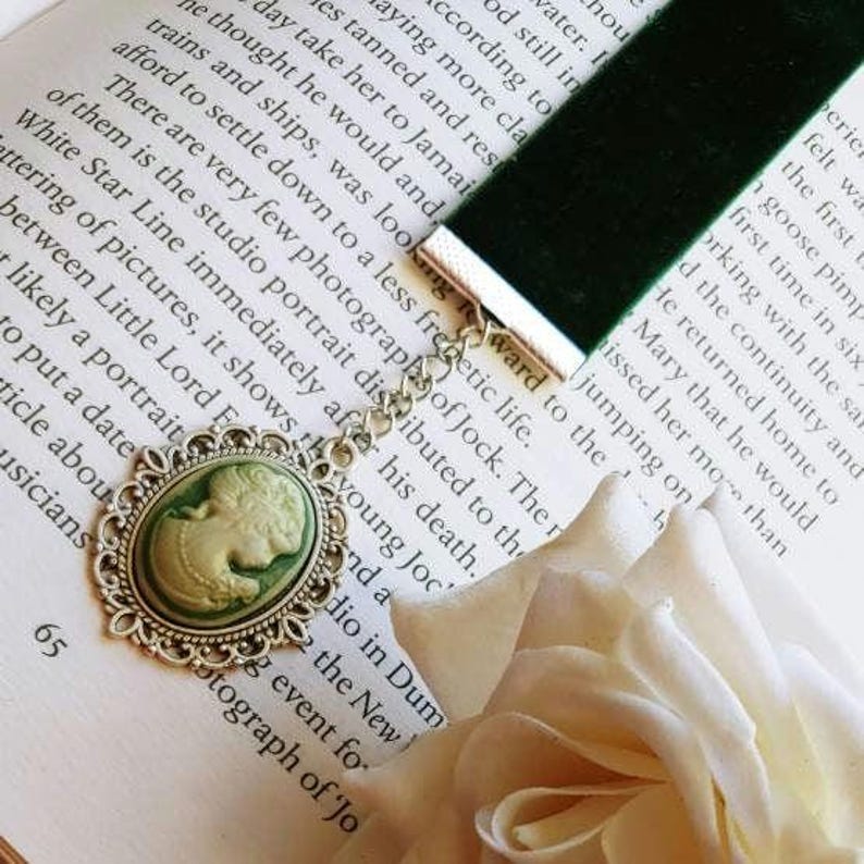 May include: A green velvet bookmark with a silver chain and a cameo pendant. The cameo is oval shaped and features a white porcelain image of a woman with a green background. The bookmark is lying on a book with a white rose.