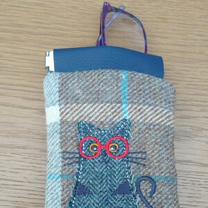 May include: A brown and blue tweed eyeglasses case with a cat design. The cat is embroidered in shades of blue and gray, wearing red glasses. The case has a blue leather top and is holding a pair of purple eyeglasses.