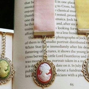 May include: Three gold-toned bookmark chains with cameo pendants. The cameos are oval-shaped and feature a white cameo profile on a green, red, and yellow background. The chains are attached to a black, pink, and yellow ribbon.