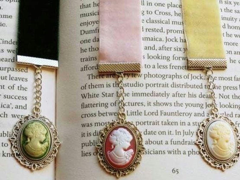 May include: Three gold-toned bookmark chains with cameo pendants. The cameos are oval-shaped and feature a white cameo profile on a green, red, and yellow background. The chains are attached to a black, pink, and yellow ribbon.