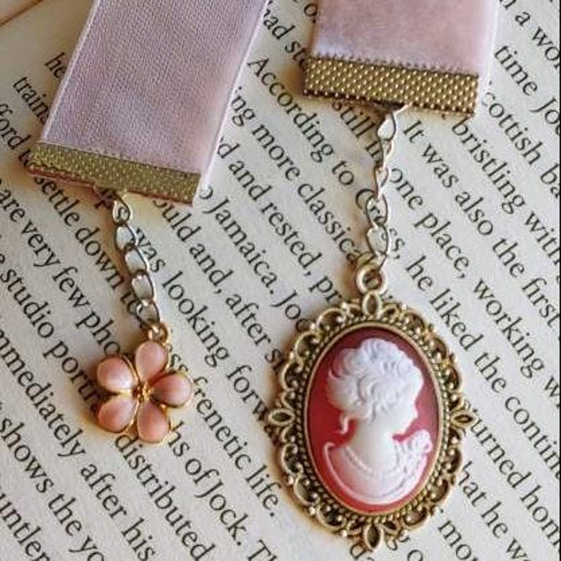 May include: A pink satin ribbon bookmark with a gold chain and a cameo charm. The cameo features a white woman with a red background. The bookmark also has a pink flower charm.