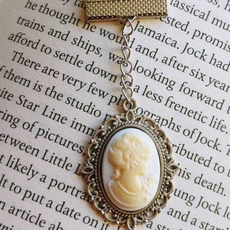 May include: A silver-toned bookmark with a cameo featuring a woman's profile. The cameo is white with a delicate floral design. The bookmark is attached to a chain with a silver-toned clasp.
